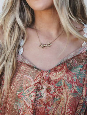 A close-up of a gold 'MAMA' letter necklace worn against sun-kissed skin, layered over a boho paisley top with lace trim peeking at the shoulder — the kind of piece that feels personal the moment you put it on.