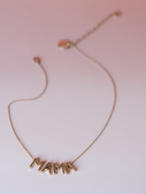 A gold 'MAMA' letter necklace with a playful bubble-font pendant, laid against a soft pink gradient background with dried floral accents — the kind of piece that feels personal before it's even gifted.