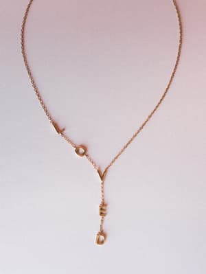 A dainty gold chain necklace spelling out 'LOVED' in spaced letter charms — part of the word runs along the chain, the rest drops in a lariat-style pendant — shot flat against a soft pink-to-white gradient background.