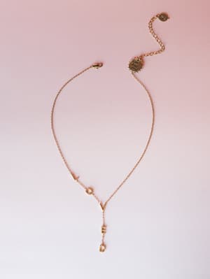 A delicate gold lariat-style necklace spells out 'LOVED' in block letter charms along the chain, photographed flat against a soft pink-to-white gradient background — the kind of piece you wear when you want a little reminder sitting close to your col
