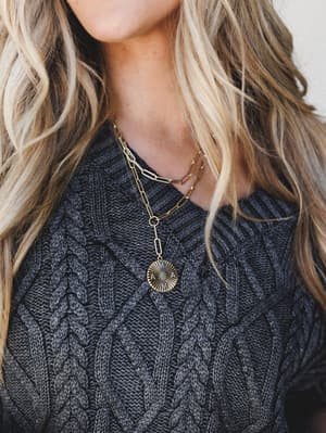 A close-up detail shot of a layered gold chain necklace with an engraved 'MAMA' sunburst medallion pendant, worn over a chunky charcoal cable-knit sweater — the kind of piece that feels personal before you even read what it says.