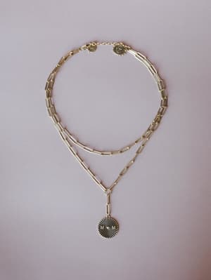 A layered gold paperclip chain necklace styled flat on a soft blush background, anchored by a circular medallion pendant engraved with 'M♥M' and a sunburst detail — the kind of piece that feels personal before you even put it on.
