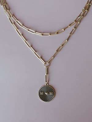 A layered gold paperclip chain necklace styled flat against a soft blush background, finished with an engraved medallion pendant featuring 'M♥M' and a sunburst pattern — the kind of piece that feels personal before you even put it on.