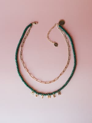 A layered necklace set styled flat on a blush pink background — a deep emerald green faceted bead strand spelling out 'MOTHER' in gold letter charms, paired with a delicate gold paperclip chain.