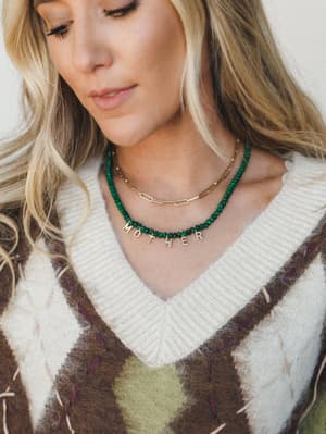 A close-up lifestyle shot showcasing a layered necklace moment — an emerald green beaded 'MOTHER' letter necklace stacked with a gold paperclip chain, worn over a cream ribbed sweater and argyle sweater vest, with a soft, intimate downward gaze.