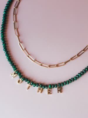 Two layered necklaces styled flat against a soft blush background — a green faceted bead strand spelling out 'MOTHER' in gold letter charms, paired with a gold paperclip chain necklace.