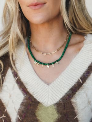A close-up shot of layered necklaces styled against a cozy argyle sweater vest — the star is a deep emerald beaded necklace spelling out 'MOTHER' in gold letters, layered with a dainty gold paperclip chain.