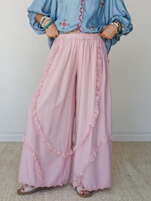 A dreamy wide-leg pant in soft blush pink with layered ruffle and lace trim panels, styled with a chambray embroidered top, stacked bead bracelets, and sandals — the kind of outfit that feels like a good mood you can wear.