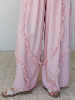 A close-up of wide-leg pink pants with delicate lace ruffle trim running diagonally across each leg, paired with white leather sandals — soft, dreamy, and the kind of thing you wear when you want the day to feel a little more special.