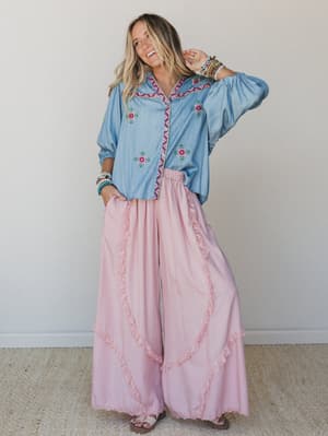 A wide-leg pink pant with ruffle and lace trim detailing is styled with an embroidered chambray button-down blouse — the whole look reads effortlessly free-spirited, like she's got somewhere fun to be and took the scenic route.