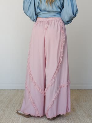 Shot from behind, a model wears ultra-wide-leg pink pants with sweeping lace ruffle trim detailing paired with a soft blue puff-sleeve top — the kind of outfit that feels like a daydream you'd actually wear to brunch.