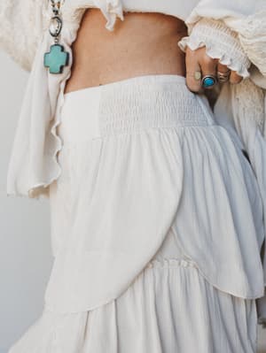 A close-up detail shot of flowy cream wide-leg pants with a smocked waistband and layered petal hem, styled with a cropped lace-trim top and stacked turquoise and silver rings — the whole look reads like a summer afternoon with nowhere to be and ever