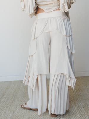 A dreamy back-facing shot of wide-leg pants layered with cascading petal-shaped ruffles in a soft natural cream — the kind of piece that makes you walk slower just to feel the movement.