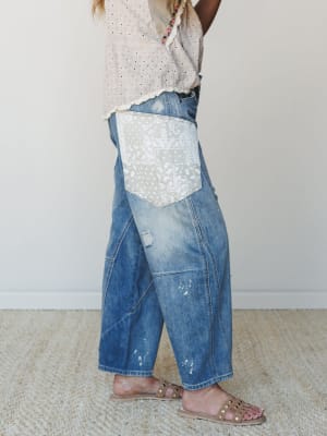 A side-profile shot of wide-leg cropped denim jeans with a standout paisley patchwork pocket, paint splatter details, and distressing — styled with a blush eyelet top and studded sandals for that perfectly lived-in, expressive feel.