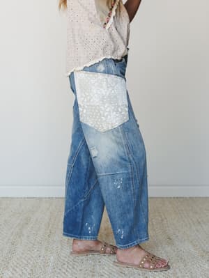 A side-profile shot of wide-leg cropped denim jeans with a standout paisley patchwork pocket, paint splatter details, and distressing — styled with a blush eyelet top and studded sandals for that perfectly lived-in, expressive feel.