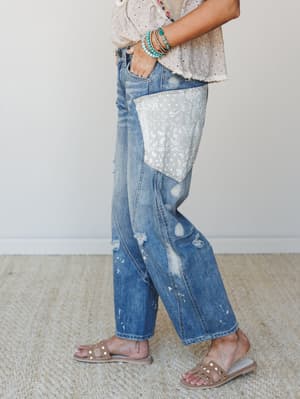 A side-profile shot of relaxed, distressed straight-leg jeans with a standout paisley patch pocket, styled with an eyelet top, stacked turquoise bracelets, and studded slide sandals — the kind of outfit that looks effortless but absolutely wasn't an