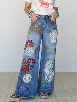 Wide-leg embellished jeans covered in crochet doily appliqués, floral embroidery, and red felt-like patchwork motifs, styled with a graphic tee and stacked bracelets — the kind of jeans that tell a whole story before you say a word.