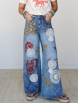 Wide-leg denim jeans covered in crochet doily appliqués, floral embroidery, and bold red folk-art motifs — styled with a graphic tee and stacked bracelets for a lived-in, expressive boho look.