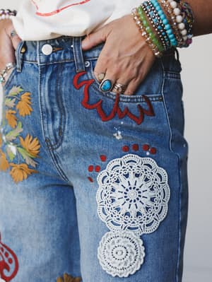 A close-up detail shot of heavily embellished denim jeans — crochet doily patches, red embroidery, and floral appliqué layered across medium-wash denim, styled with a stack of beaded bracelets, turquoise rings, and a white graphic tee peeking in at t