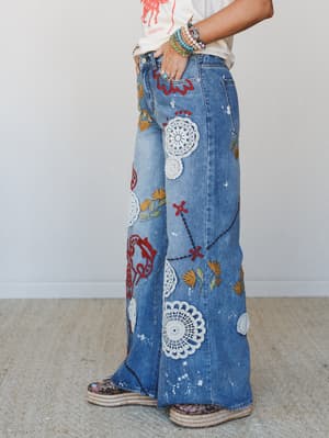 Wide-leg denim jeans covered in doily appliqués, red embroidery, floral stitching, and paint splatter — styled with a graphic tee, stacked bracelets, and espadrille platforms for a look that feels like a vintage treasure hunt paid off.