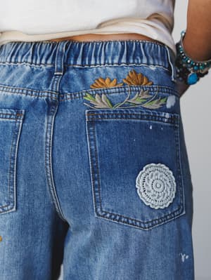 A close-up detail shot of medium-wash denim jeans with floral embroidery above the back pocket and a crochet medallion patch — the kind of jeans that make people ask where you got them at the farmers market.