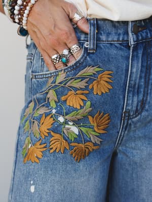 A close-up detail shot of medium-wash distressed denim jeans featuring large-scale floral embroidery in golden amber and sage green across the hip and thigh, styled with layered silver rings, a turquoise stone ring, and stacked beaded bracelets.