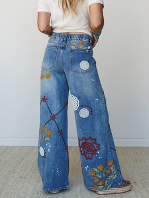 A model shot from behind showcasing wide-leg denim jeans covered in floral embroidery, crochet doily appliqués, and hand-stitched detailing — paired with a white crop tee and espadrille wedges for an effortlessly boho, lived-in feel.