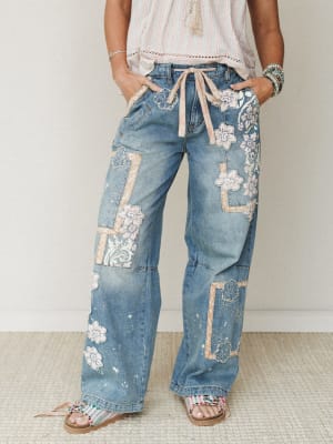 Wide-leg patchwork denim jeans covered in floral appliqués and lace-edged fabric patches, styled with a striped top, beaded bracelets, and colorful sandals — the kind of jeans that do all the talking.