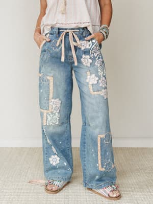 Wide-leg patchwork denim jeans covered in floral appliqués and lace patches, styled with a striped crop top and beaded sandals — the kind of jeans that have a whole personality before you even put them on.