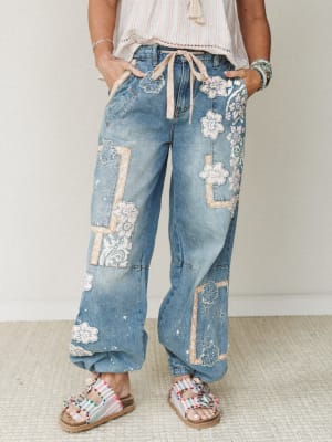 Wide-leg patchwork jeans covered in floral appliqués and lace panels styled with a striped top, stacked bracelets, and beaded sandals — the kind of jeans that make a plain white tee feel like a whole personality.