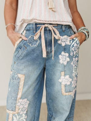 A close-up of wide-leg denim jeans covered in floral appliqué patches and embroidery, tied at the waist with a soft ribbon — styled with a striped top, layered bracelets, and a tassel necklace for an effortlessly boho feel.