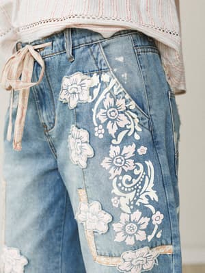 A close-up detail shot of light-wash denim jeans covered in white floral appliqué patches and painted floral scroll work, paired with a striped lace-trim top with a trailing tie — the kind of jeans that make people stop and ask where you got them.