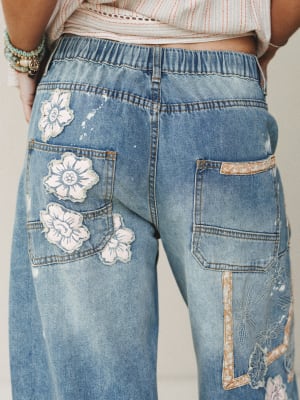 A close-up back view of light wash wide-leg jeans featuring floral appliqué patches and embroidered botanical details — the kind of jeans that feel like a whole personality in denim form.