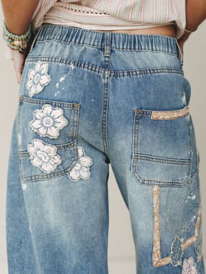 A close-up back view of light wash wide-leg jeans featuring floral appliqué patches and embroidered botanical details — the kind of jeans that feel like a whole personality in denim form.