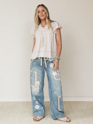 A breezy full-body shot of a model in wide-leg patchwork denim jeans and a flowy striped ruffle-sleeve top, styled with layered boho jewelry against a soft neutral studio wall — the kind of outfit that looks effortless but was definitely thought abou