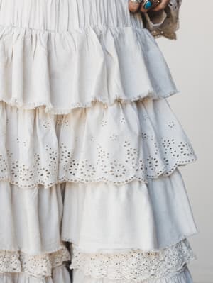 A close-up detail shot of multi-tiered ruffle pants in cream and soft grey, layered with eyelet trim and raw frayed edges — the kind of piece that looks like it has a whole story already.
