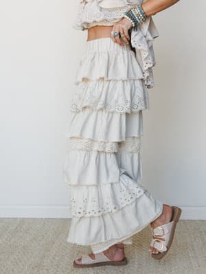 A dreamy tiered ruffle maxi skirt or wide-leg pant in soft taupe, layered with eyelet lace trim throughout — styled with stacked turquoise bracelets, silver rings, and neutral buckle sandals for an effortlessly boho look.