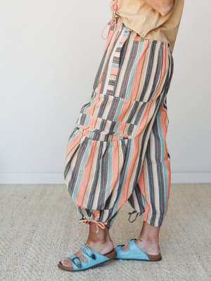 A side-angle shot of voluminous striped harem pants in a warm multicolor stripe — paired with a sandy linen top and sky blue sandals for an effortlessly earthy, well-traveled feel.
