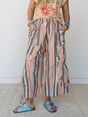 A cropped wide-leg pant in a bold multicolor vertical stripe steals the show, styled casually with a vintage graphic tee, stacked turquoise bracelets, and blue footbed sandals — effortless and a little retro in the best way.