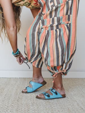 A close-up lifestyle shot of relaxed striped harem pants with drawstring ankles, styled with sky blue buckle sandals and stacked turquoise bracelets — the kind of outfit that says 'I know exactly what I'm doing' without trying.