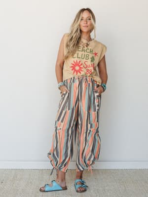 A breezy full-body model shot of multicolor striped wide-leg pants paired with a sandy graphic tee and turquoise sandals — the kind of outfit that looks like it was pulled together effortlessly somewhere between a farmer's market and a beach town.