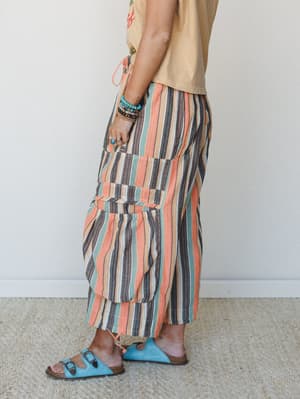 A side-profile shot of wide-leg striped pants in warm coral, sage, and charcoal tones, styled with a sandy graphic tee, turquoise sandals, and stacked boho jewelry — the whole look has that easy, unhurried summer energy.