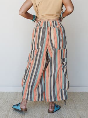 A back-view shot of wide-leg striped pants in a warm sunset palette — coral, sage, tan, and charcoal — styled with a cropped mustard top, turquoise beaded bracelets, and teal tassel mules for a relaxed, well-traveled feel.