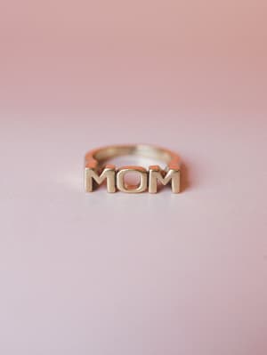 A gold 'MOM' letter ring shot up close against a soft blush pink gradient background — simple, sentimental, and the kind of thing you'd want to wear every single day.