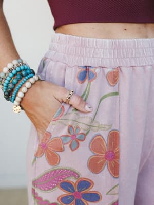 A close-up detail shot of lavender floral wide-leg pants with a smocked waistband and side pocket, styled with a wine crop top and a stack of beaded bracelets — the gold 'MOM' ring on her finger is the quiet little centerpiece.