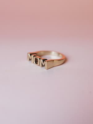 A gold 'MOM' letter ring shot solo against a soft pink gradient backdrop — the kind of piece that's small but says everything.