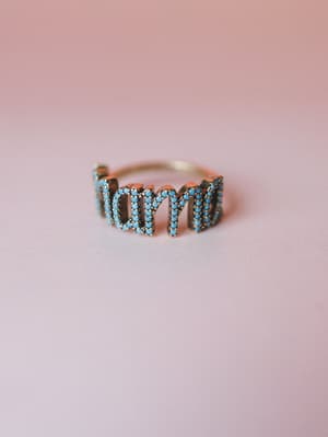 A close-up product shot of a word ring spelling out 'mama' in turquoise stone-set lettering on an antique gold band, photographed against a soft dusty pink surface — the kind of piece that feels personal before you even put it on.