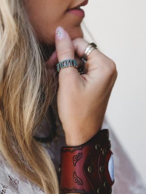 A close-up lifestyle shot of stacked rings and a wide tooled leather cuff on a model with blonde hair — the turquoise 'mama' letter ring is front and center, layered with a silver band, and the whole moment feels personal, a little worn-in, and very