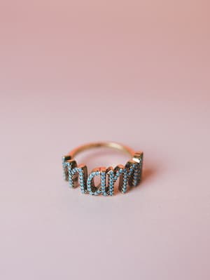 A gold 'mom' letter ring set with turquoise stones, photographed up close against a soft pink background — the kind of piece you wear every day and never take off.