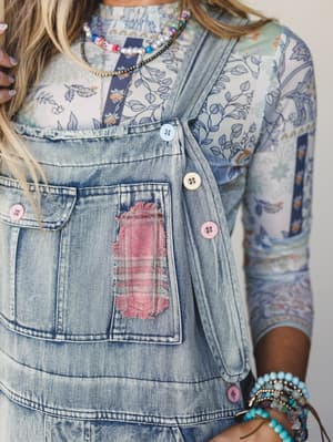 A close-up detail shot of light-wash denim overalls layered over a mixed floral print long-sleeve top, styled with stacked beaded necklaces and turquoise bracelets — the kind of outfit that looks effortless but definitely took thought.